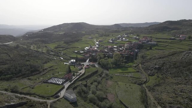 Smooth aerial flyover above Albergaria da Serra revealing traditional Portuguese architecture, cultivated fields, and the natural beauty of Serra da Freita. Drone rotating to the left.