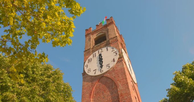 Belvedere Tower in Mondov&igrave; rises above green trees, its large white clock face and Italian flag lit by bright daytime sun.