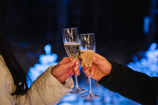 Romantic winter toast with champagne glasses, celebrating a marriage proposal or Valentine's Day. A couple holding sparkling drinks on a snowy terrace at night.