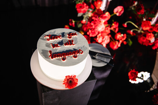 Top down view of a minimalist wedding cake with the romantic inscription "Love you in all languages", showcased on a pedestal with vibrant red floral decor in the background.