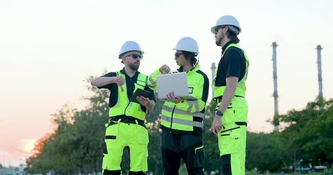 Male engineer speaks through walkietalkie while refinery technician reviews laptop and tablet, managing pipeline inspection workflow, emergency communication, maintenance coordination onsite.