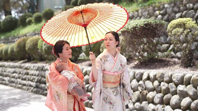 Three women in traditional kimonos walk gracefully. One carries a large, ornate paper parasol. They stroll beside a stone-lined garden path. Their expressions convey calm, gentle joy