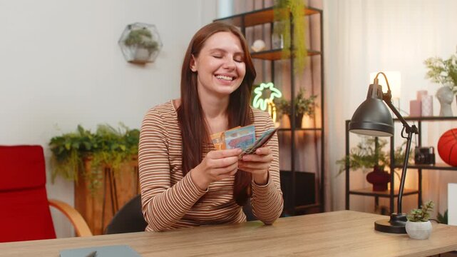 Young woman at home counts cash euro bills stacks money and smiles pleased about salary bonus or wins. Caucasian girl at desk checks amount puts bills in wallet and notes budget plan with calm focus