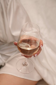 Glass with wine. Closeup of a woman's hand holding a glass of white wine. She wears a white silk robe and has a french manicure. The setting is indoors, during a wedding morning.