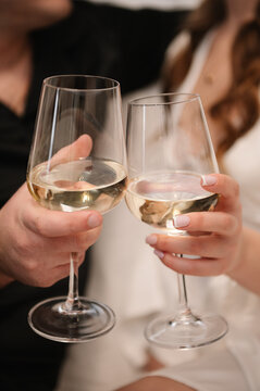 Toast closeup. Cheers. A romantic close-up of a man's and a woman's hands clinking glasses of white wine. The setting is indoors, during a wedding morning or celebration. Soft focus background.
