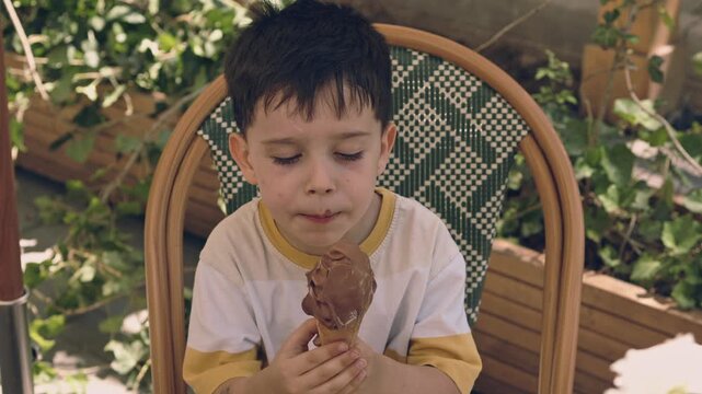 kid, boy, sitting on a woven chair outdoors and eating chocolate ice cream from a waffle cone. The child is holding the dessert with both hands during a sunny summer day.