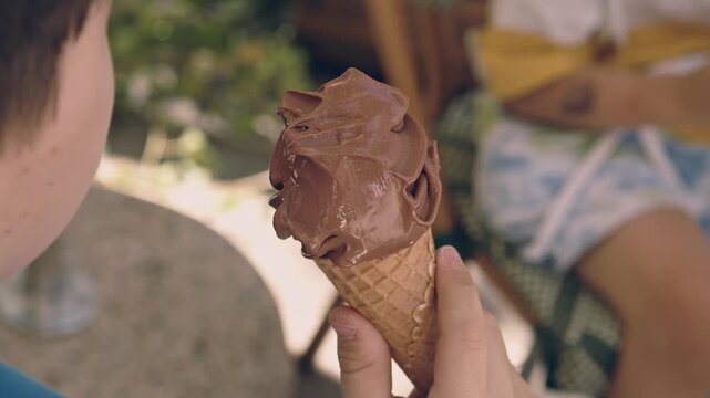 kid, boy, sitting on a woven chair outdoors and eating chocolate ice cream from a waffle cone. The child is holding the dessert with both hands during a sunny summer day.
