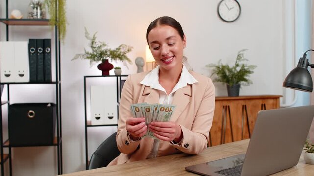 Young businesswoman at home office holds dollars counts cash and checks total smiles at strong profit. Freelancer girl at table writes note for revenue adds stack to safe envelope and plans next save