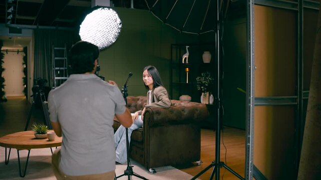 Young female asian influencer sitting on a sofa while a cameraman is filming her. Professional studio setup with lights and microphone
