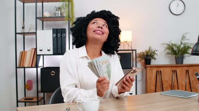 African American businesswoman at home office laughs holding smartphone surprised by profit alert counts dollar cash. Freelancer girl at table plans spending smiling imagining travel fun retail dreams
