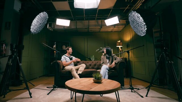 Cheerful male and female hosts recording an audio podcast. Modern interview setup with professional microphones, cameras, and lighting