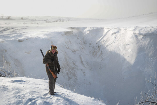 A hunter in warm winter clothes and a fur hat stands on the edge of a deep snow-covered ravine.