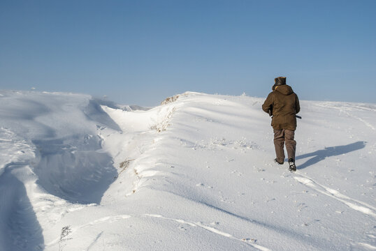 A hunter in warm winter clothes walks along a snow-covered field along a ravine. Back view.