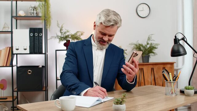 Middle-aged businessman at home office scrolls smartphone for fresh inspiration then grabs pen writing notes. Freelancer guy at table concentrates handwriting solution into notebook staying focused.