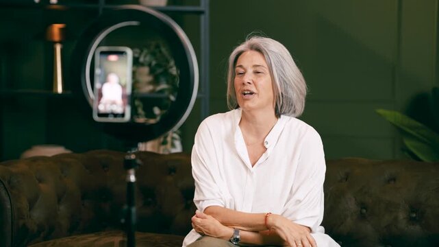 Mature woman with grey hair talking to her smartphone camera on a tripod with a ring light. Female influencer recording a video for her channel
