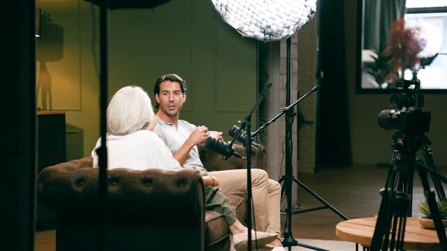 Young professional photographer being interviewed by a senior host during a podcast show in a modern recording studio with video equipment