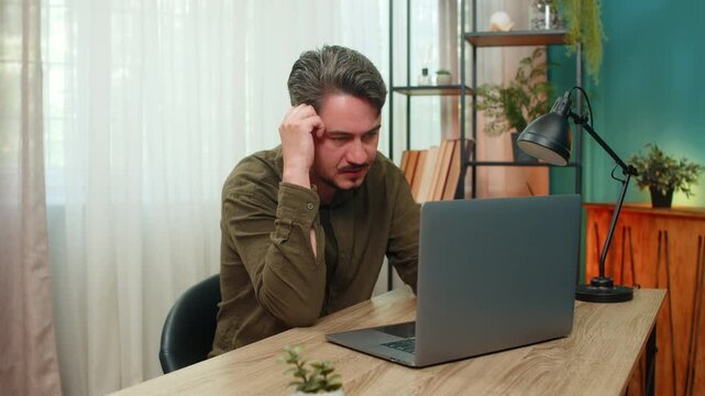 Middle-aged man at home works on laptop frowns as error message shows bad news virus warning causing stress. Young adult guy at table tries fix computer issue feeling tense urgent pressure frustrated