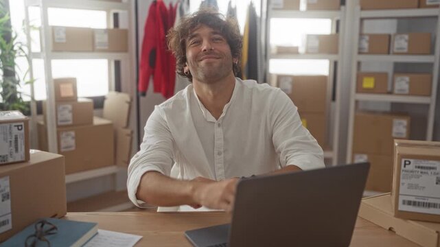 Man smiling with arms crossed at desk with laptop and stacked shipping box in building; optimism entrepreneurship.