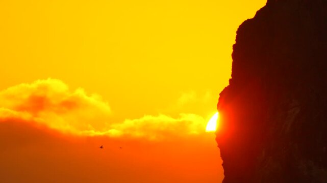 Sunset rock sky dramatic sun setting behind a massive coastal cliff silhouette with birds flying through the orange clouds