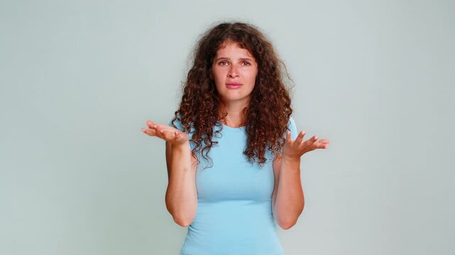 Young woman holding hand near ear gesture indicating difficulty hearing clearly, confused emotional reaction. Girl on gray background visibly expresses uncertainty asking to speak louder humorous mood