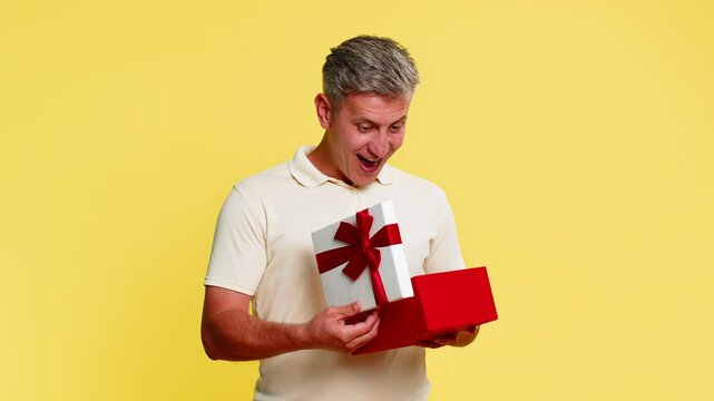 Middle-aged man opening gift box and suddenly freezing with amazed expression and bright joyful face reacting to unexpected pleasant surprise. Young guy showing happiness isolated on yellow background
