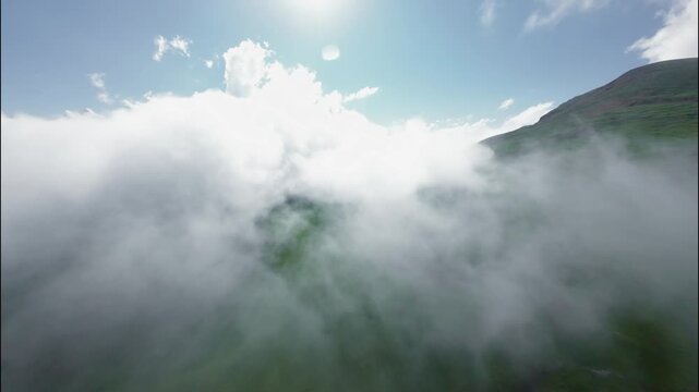 Aerial view of clouds rolling over green hills under bright blue sky. View from FPV drone.
