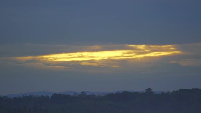 Close-up captures the morning sky in Banten as the sun rises behind heavy clouds. Golden light fringes the vaporous edges above dark silhouetted trees during the early dawn hours. 