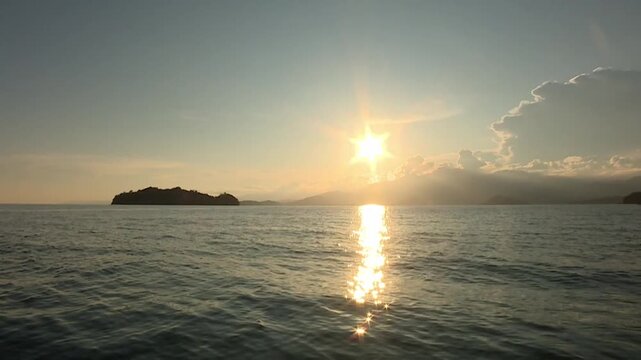 Wide shot captures the sun descending toward the western horizon over Central Java waters. Sunlight reflects on the ocean surface, creating sparkling glimmers and star-like patterns on the sea. 