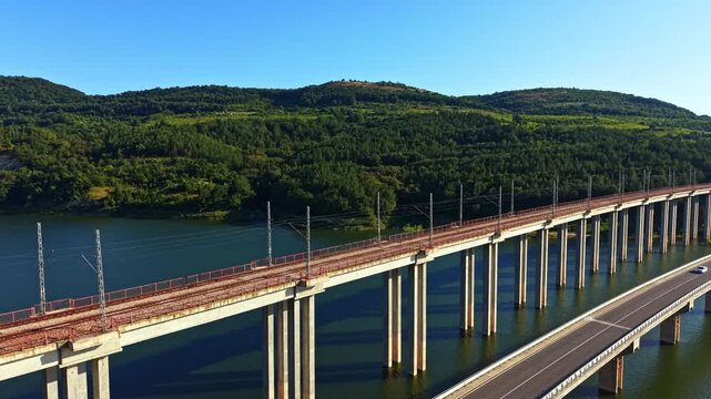 A train bridge spans over a river with a road running alongside it. Green hills rise in the background under clear blue sky. The scene shows a typical day with no people in sight.