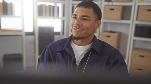 Young hispanic man with smiling face, wearing navy jacket and white tee, looking aside in an office building; quiet confidence.