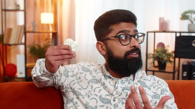 Indian young man sitting at home on sofa, holding two golden bitcoin BTC coins and seriously thinking about crypto investment future. Arabian guy analyzing financial risks and market direction indoors