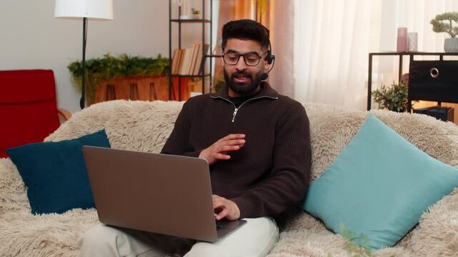 Indian man at home speaks on laptop video call using wireless headset, gesturing while talking with client. Arabian guy on sofa works in call center remotely, staying focused and confident online.