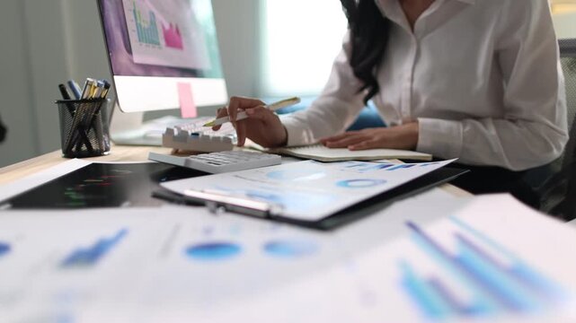 Businesswoman is working at her desk, analyzing financial data and taking notes. She is using a calculator, a pen, and a notebook.