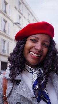 Vertical. African millennial woman walks along a Paris street wearing a red beret and coat, smiling with direct gaze amid Haussmann buildings, urban travel mood on a cloudy day.