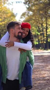 vertical. Interracial couple piggyback walking an autumn park, smiling and holding hands. Travel portrait in natural light with leaves and trees, conveying romance, connection and joyful lifestyle