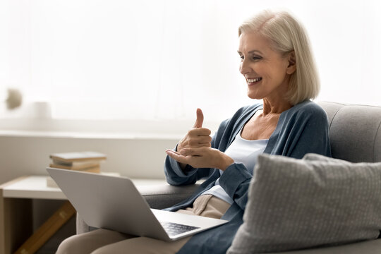 Perfect choice. Smiling elderly woman blogger influencer sit on couch show thumb up to laptop webcam advice audience to vote for post recommend goods service. Happy old lady leave good feedback online