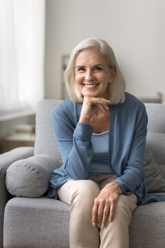 Age does not matter. Happy carefree senior adult woman touch chin with smile look at camera sit on comfy sofa. Smiling elegant mature female retiree spend free time at home enjoy good day perfect mood