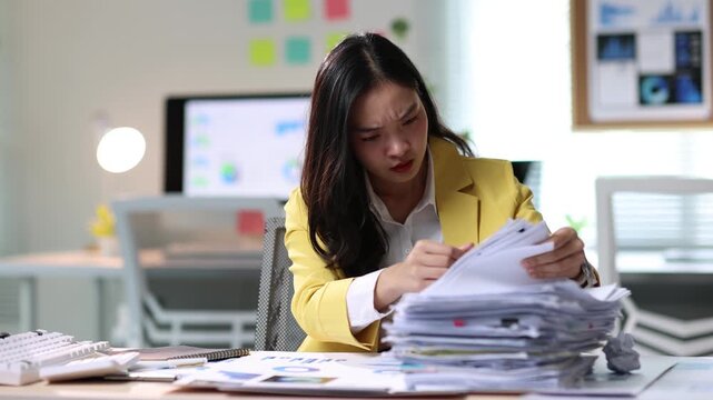 Office worker feeling stressed working with documents. Asian office worker sits at her desk, overwhelmed by a mountain of paperwork. She stressed as she tries to manage the workload.
