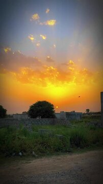 A beautiful golden sunset illuminates the sky with glowing orange clouds over a peaceful rural landscape, green fields, trees, and distant buildings. 