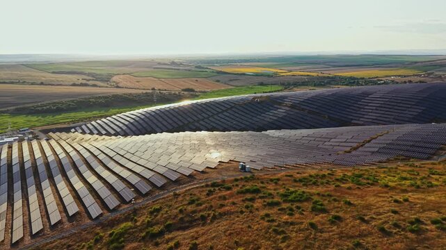 Rows of solar panels fill the landscape in a rural setting. The sun shines brightly as workers tend to the installation. Green fields stretch in the background under a blue sky.