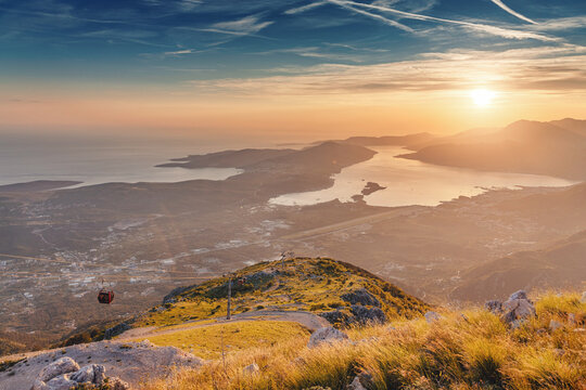 Panoramic aerial view showing a cable car ascending a mountain with the Bay of Kotor and Adriatic Sea glowing under a sunset sky