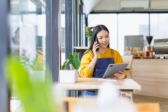 Asian woman cafe owner managing small business, talking on smartphone and checking orders on tablet