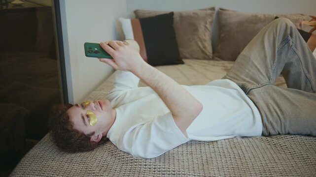 Person lying on a bed looking at a smartphone while wearing eye patches in a cozy indoor space during the day