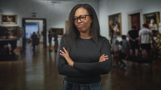 Woman wearing glasses with arms crossed in museum building among paintings; confidence self reflection.