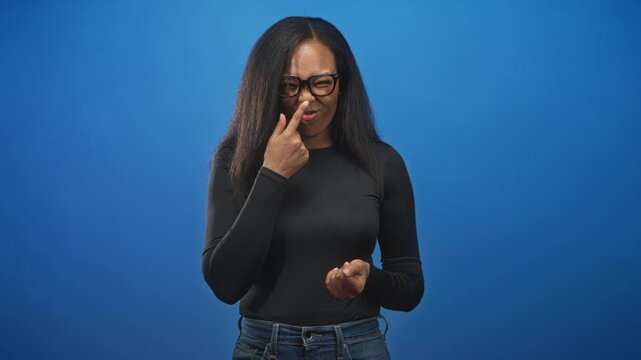 Young african american woman wearing glasses and a black top pinches her nose, gestures with both hands and grimaces against a blue studio backdrop; disgust discomfort.