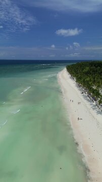 Vertical drone flies over and ascends along Dumaluan Beach, showing white sand, clear shallows, swimmers, and palm-lined coast