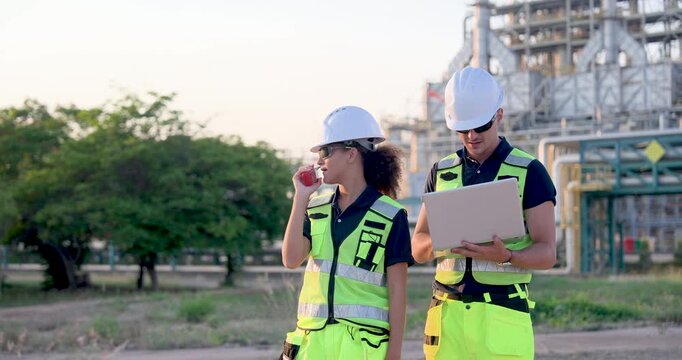 Female refinery technician reporting operational status through walkietalkie beside male engineer reviewing laptop data during petrochemical maintenance inspection near industrial pipeline system