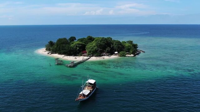Pedestal drone shot rises as a traditional pinisi boat sails away from a small Sulawesi island. The white sand shore and deep blue ocean create a stunning, visually pleasing tropical vista. 