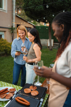 Diverse women friends enjoying backyard barbecue and using smartphone