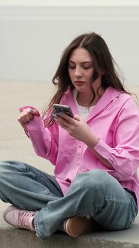 Young woman in a pink shirt sits cross legged outdoors and reads something on her smartphone with a worried thoughtful expression, reflecting digital anxiety and online communication stress.